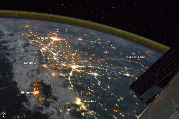 Nighttime satellite photograph showing the Indo-Gangetic Plain with visible city lights marking New Delhi, Lahore, Islamabad, and Srinagar, and a portion of the Earth's curvature with a visible atmosphere and space. An object resembling a solar panel from a satellite or space station is visible on the right side.