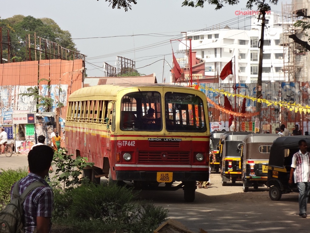 A red and yellow KSRTC ordinary bus on a street with several people around and auto-rickshaws parked nearby